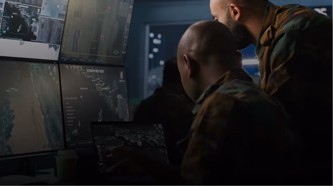 Two men in camouflage reviewing digital maps and data on multiple screens and a laptop in a dimly lit room.